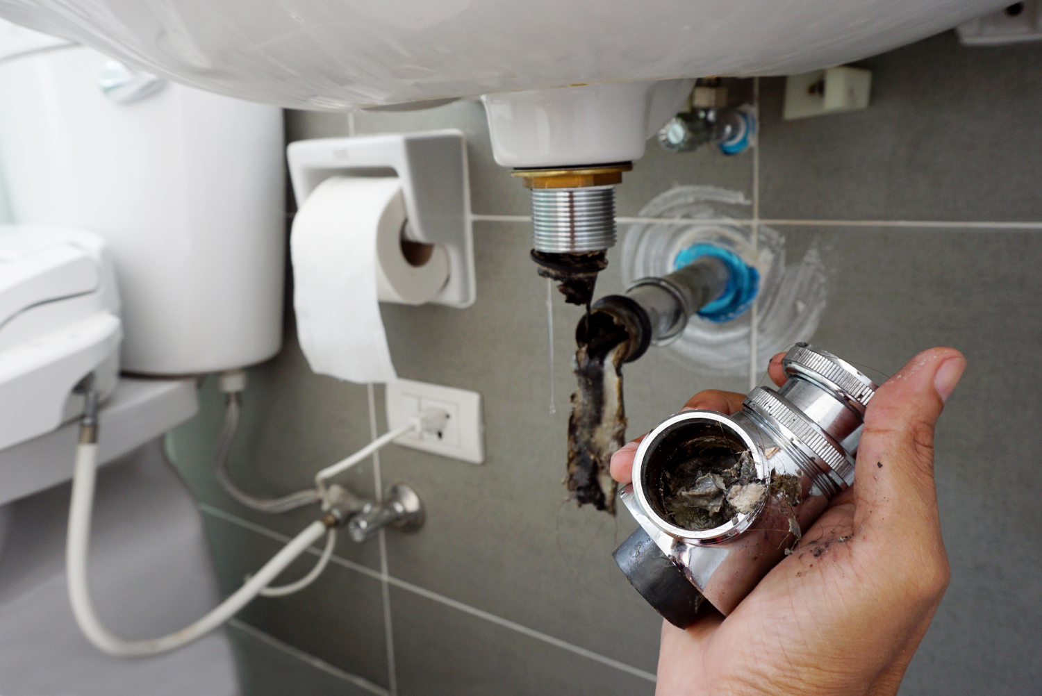 Hand holding a clogged plumbing fitting with debris, showcasing drain cleaning in a bathroom setting, relevant to Royal T Plumbing's drain cleaning services in Vancouver.