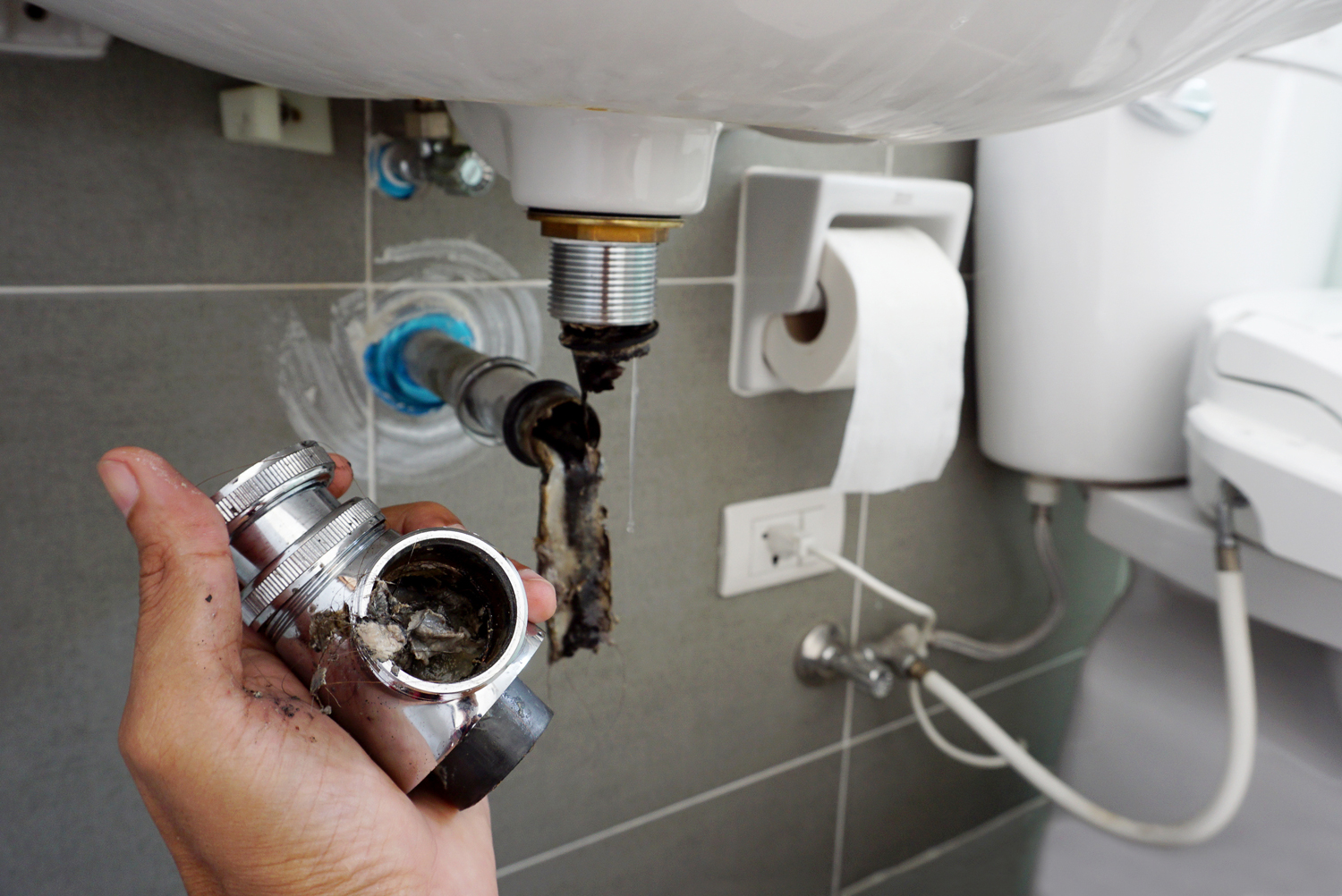 Hand holding a clogged plumbing fitting with debris, showing plumbing installation under a sink, highlighting drain cleaning service.