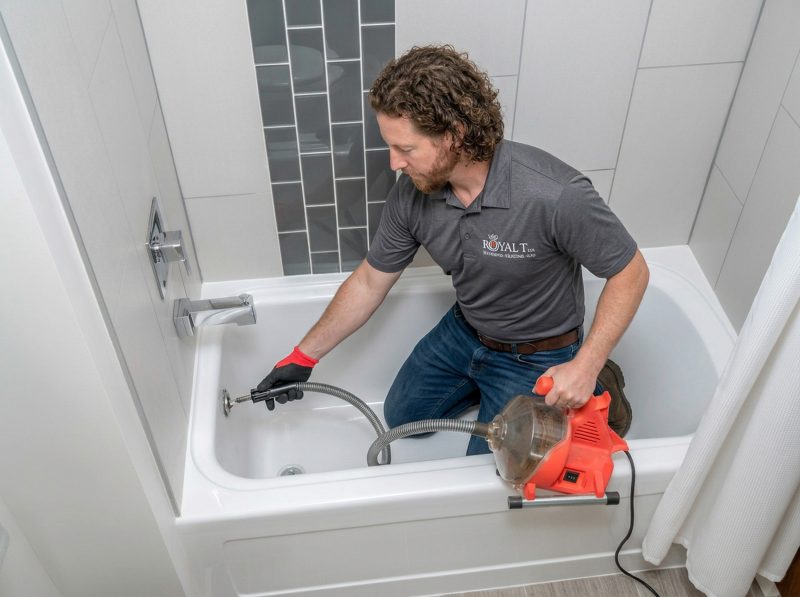 Plumber from Royal T Plumbing using a drain cleaning machine in a bathtub, addressing clogged drain issues in a residential setting.